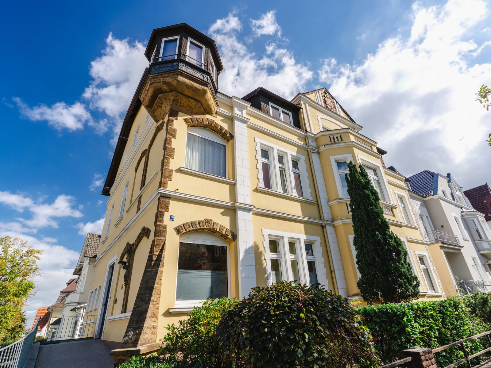 Ein imposantes, historisches Gebäude. Die Fassade ist gelb mit weißen Elementen, zusätzlich existieren Verzierungen aus Sandstein. Zu sehen ist der Gebäudeeingang auf der linken Seite, um das Gebäude herum stehen einige Bäume und Sträucher.