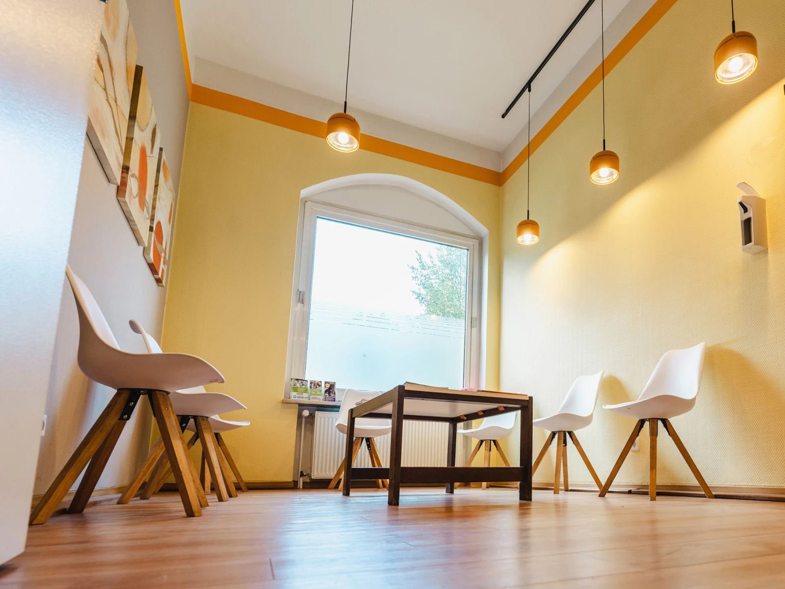 Ein Wartezimmer. Die Wände sind gelb und weiß angestrichen, und mit Bildern dekoriert. Von der Decke hängen Pendelleuchten. Um einen flachen Tisch herum stehen mehrere Stühle.